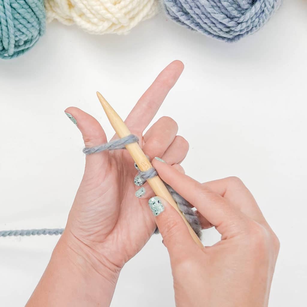The right hand guides the knitting needle under the yarn, to the base of the thumb and then up, through the loop on the thumb.