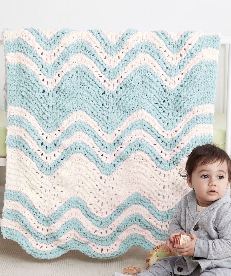 Baby Blanket Knit Patterns - baby sitting in front of a blue and white Garter Ripple Striped baby blanket draped over a crib