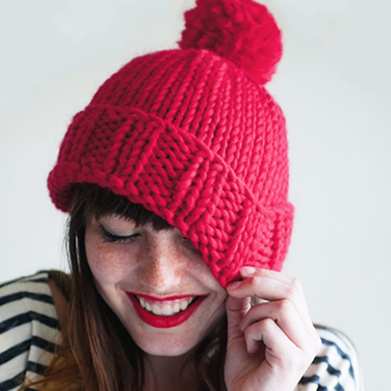 A smiling woman holding a chunky knit beanie with a pompom on top.