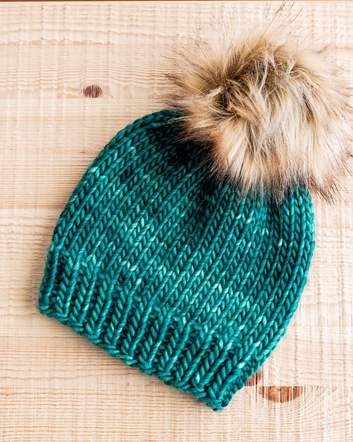 This is a photo of a knitted beanie with smooth stitches and a faux fur pompom on top.