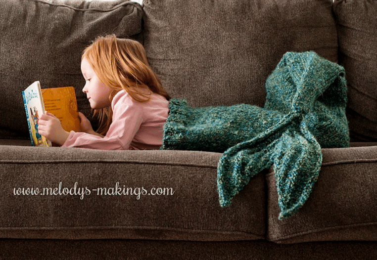 This is an image of a child reading and wearing a knit blanket that is in the shape of a mermaid tail or fish tail. This pattern shows how to knit a blanket for mermaid lovers.