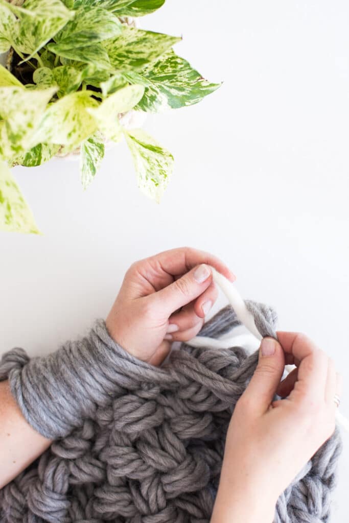 A strand of scrap yarn getting woven through chunky knit hoops. This image demonstrates how to stop in the middle of an arm knitting project.