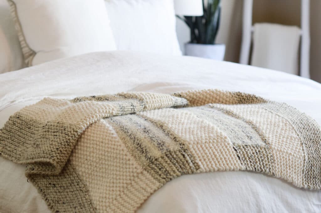 This is an image of a knit blanket draped on top of a white bed to show off the stitch patterns.