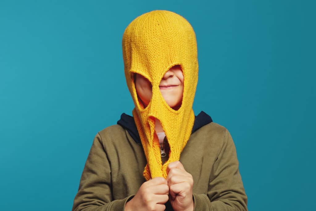 Image of a cute smiling child stretching a balaclava in front of their face. This image is intended to show off the finished item of a free balaclava knitting pattern.