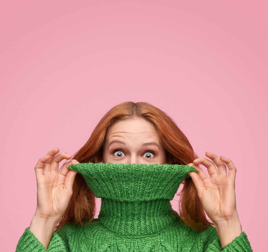 A woman stretching out a turtle neck sweater while looking at the camera. This image shows how Jeny's surprisingly stretchy bind off can look on a knitted sweater.