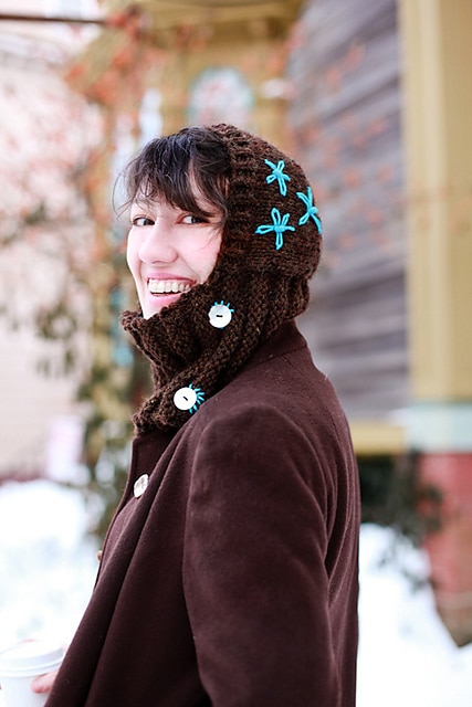 A smiling woman wearing the finished free knit balaclava pattern: Embroidered Hood. 
This is a snug balaclava with embroidery around the buttons and embroidered flowers on the hood.