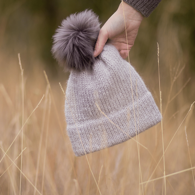 A photo of a hand holding a knit beanie with a faux fur pompom on top.