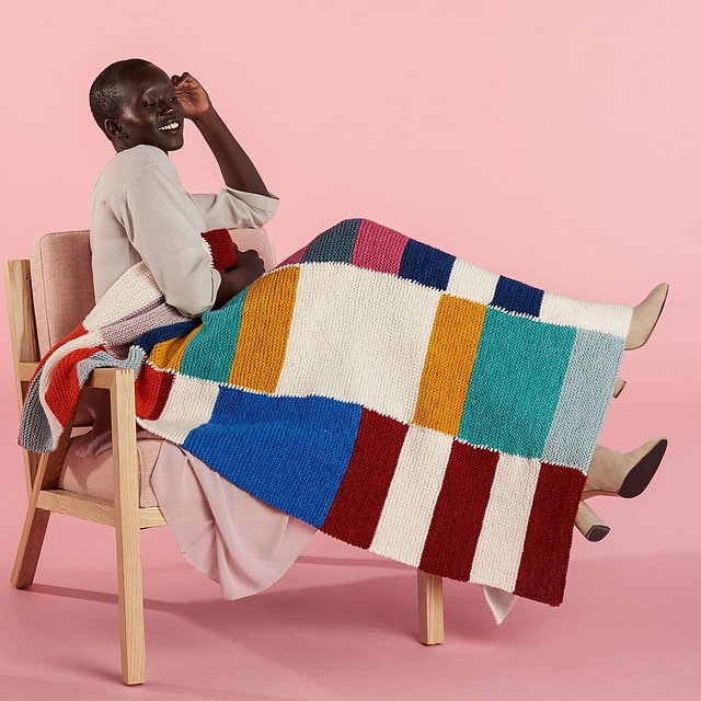 This is an image of a woman sitting and smiling on a chair with a knitted blanket draped over her legs.