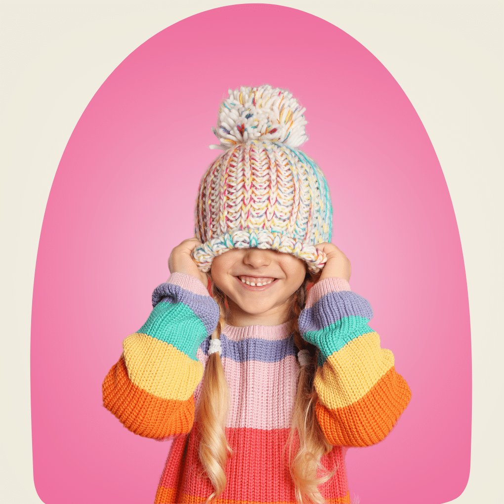 A cute, smiling child holding a colorful knitted beanie over her eyes.