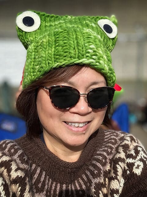 A woman smiling at the camera wearing a green knit frog hat