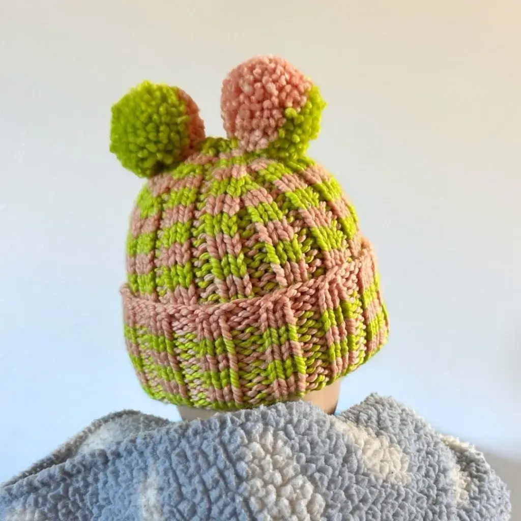 A child wearing a green and pink knit ribbed beanie with two pom poms that look like ears