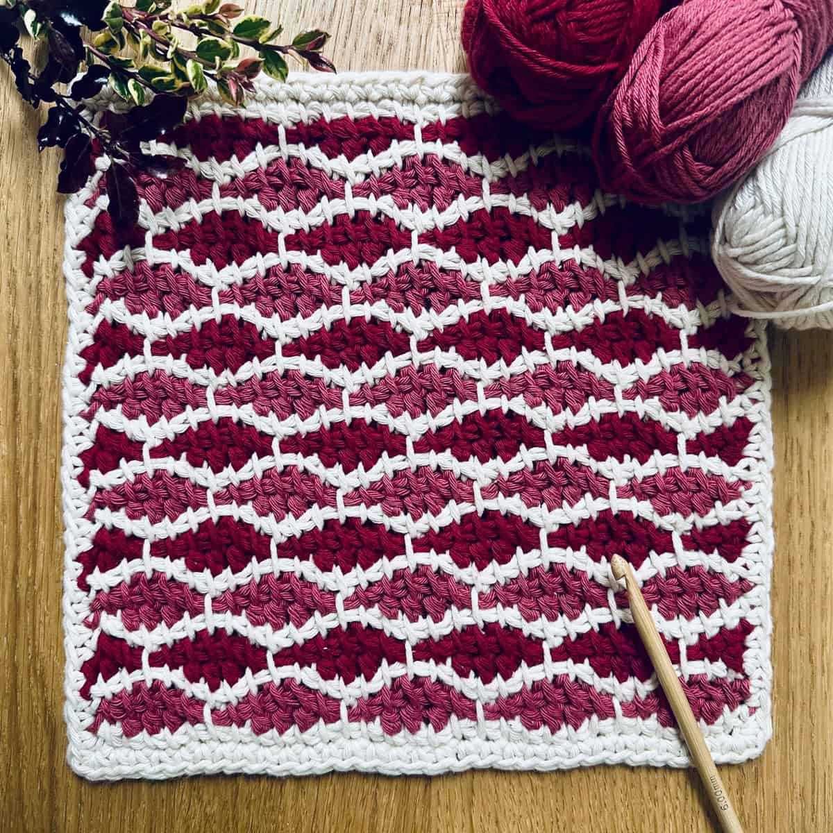 A square, crochet washcloth that has a wavy stitch design in red, pink, and white.