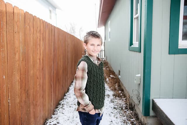 A young boy wearing a green hand knit cabled vest.