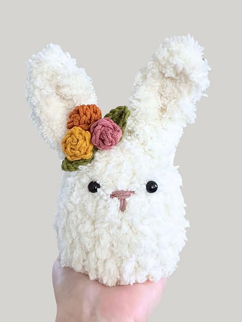 A fluffy white crochet bunny pattern with 3 small crochet rosettes by their left ear.
