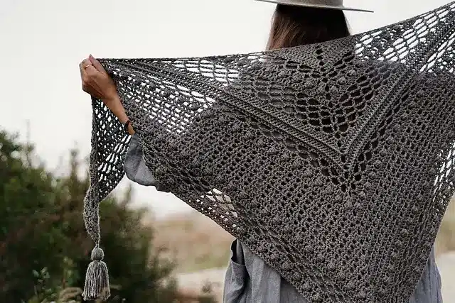 A grey textured, airy crochet shawl being draped over a woman's shoulder.