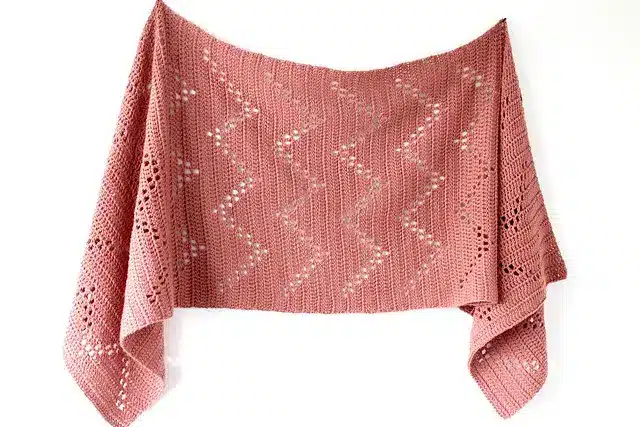 A salmon pink crocheted shawl with a zig zag or chevron filet crochet patterns running across.