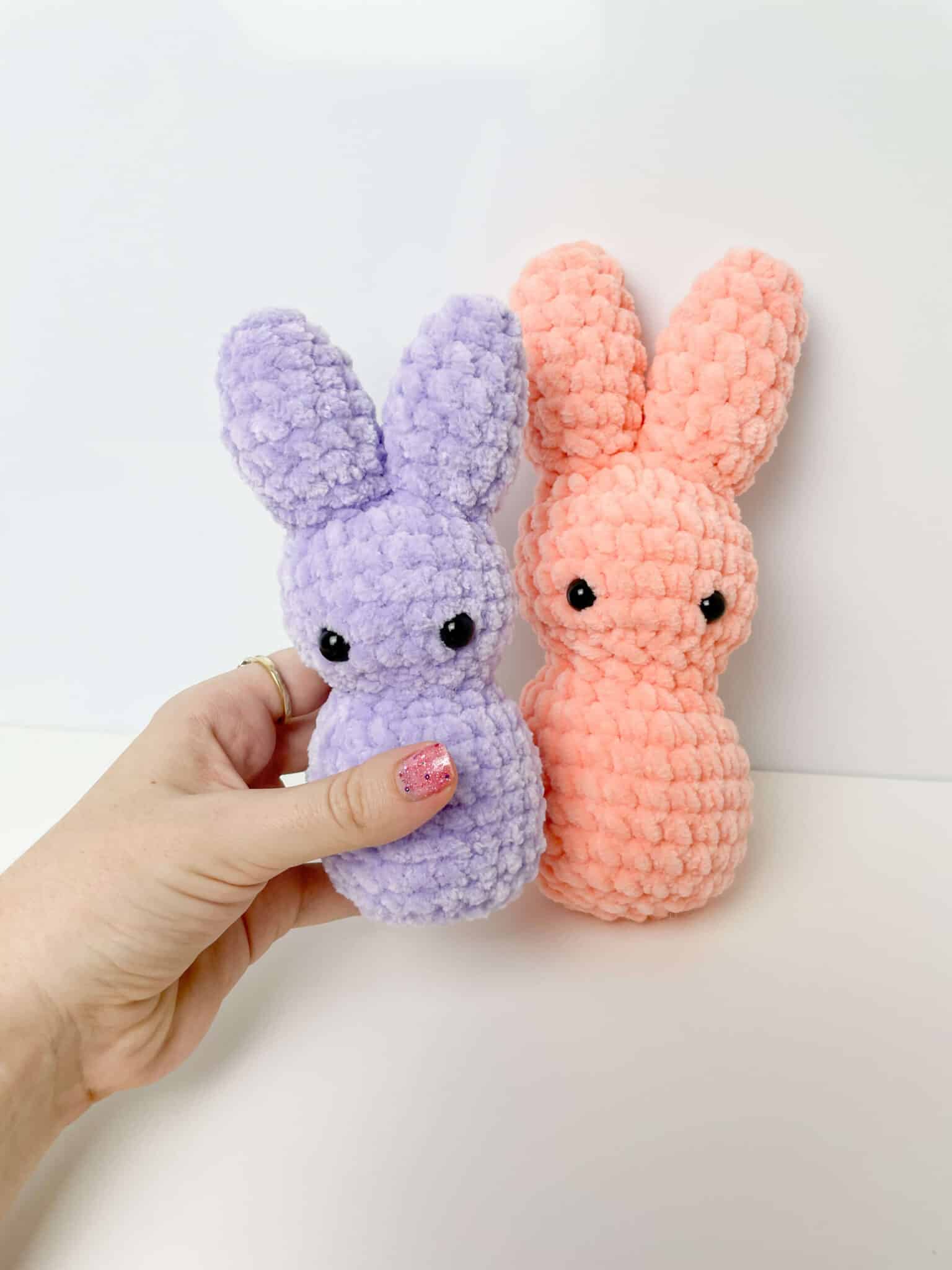 Cute amigurumi bunny peeps made with velvet yarn. The one to the left is being held by a hand is made with purple yarn and a pink bunny leaning against the wall to the right.