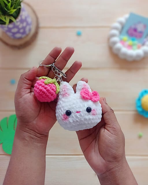 Cute white crochet bunny pattern keychain with a pink strawberry being held up by two hands.