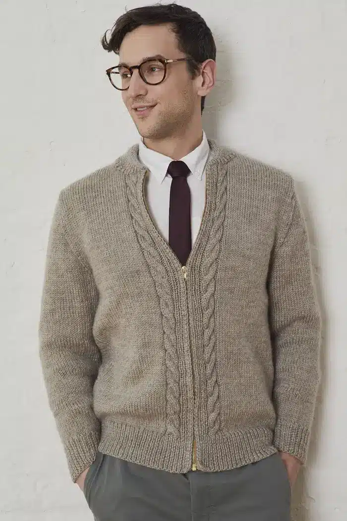 A man leaning against a wall wearing a handknit sweater with knit cables running along either side of the zipper. 