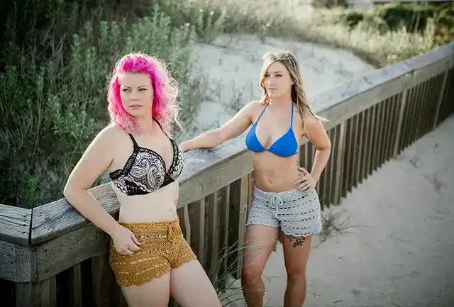 Two women wearing crochet shorts, leaning against a wooden railing at the beach.