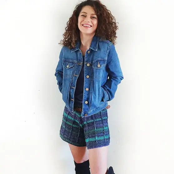 A smiling woman with curly hair, wearing a denim jacket and denim blue crochet shorts.