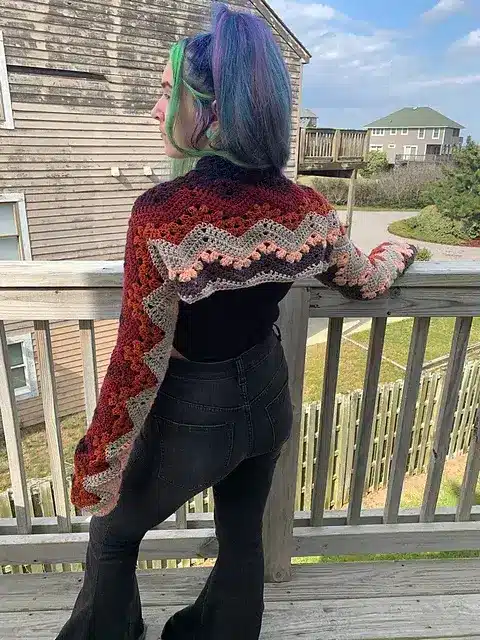 A young woman with her back to the camera, wearing a very colorful crochet shrug that has a textured, zigzag design.