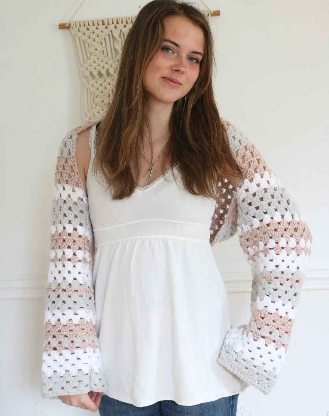 A young woman wearing a loose, white tank top, and a crochet granny square shrug.
