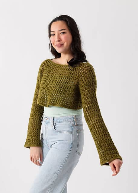 A young woman with dark hair, wearing a moss green crochet shrug over a light green tank top and blue, denim jeans.