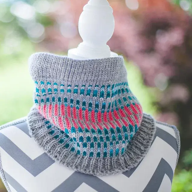 Stunning loom knit cowl wrapped around a white mannequin. It has re, blue, and grey vertical designs.