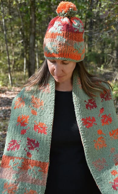 A woman wearing a stunning, intricate loom knit scarf and hat pattern set in the woods. Both the hat and scarf have autumn leaves in orange, red, pink, and yellow hues scattered all over the fabrics.