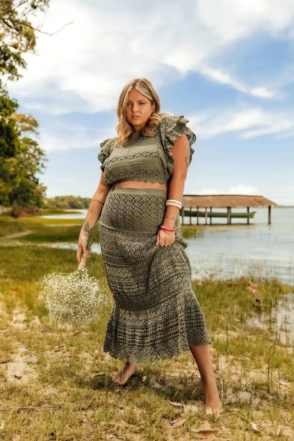A woman wearing a stunning, intricate crochet skirt made with thin, fingering yarn posing in front of a lake.