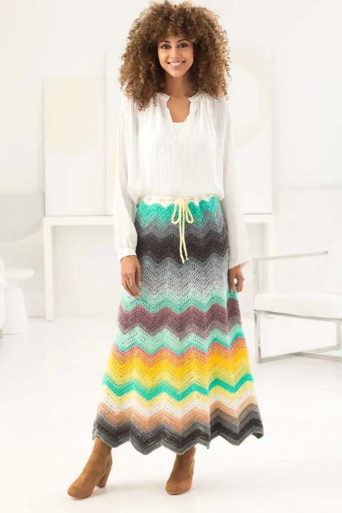 Tall woman wearing a loose, white top and a colorful crocheted maxi skirt. The skirt is made with a beautiful blend of grey, black, blue, salmon pink, yellow, and orange yarn.